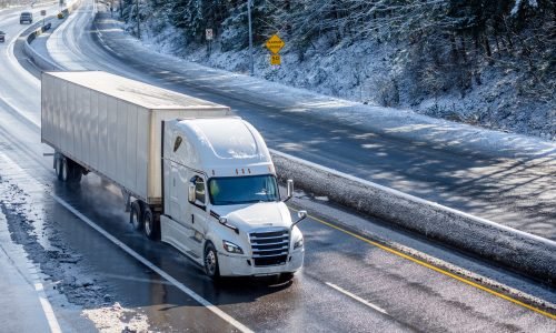 Big rig long haul gray semi truck tractor transporting commercial cargo in dry van semi trailer going on the wet slippery road with water from melting snow and winter snowy trees on the hills